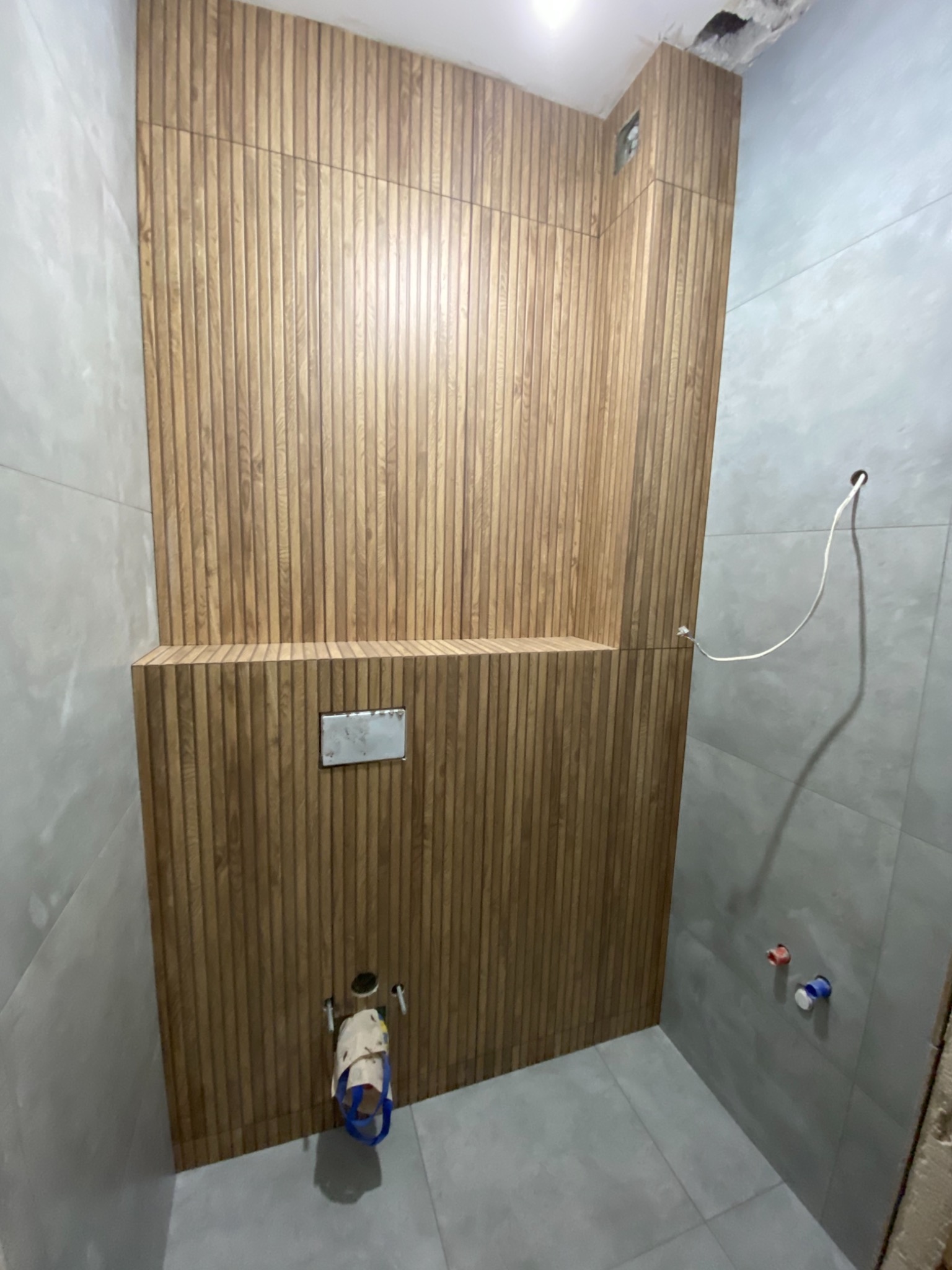 Łazienka w trakcie wykańczania: drewniane panele ścienne tworzące efekt lameli, szare płytki, wystające rury i przewody, widoczne niedokończone prace.