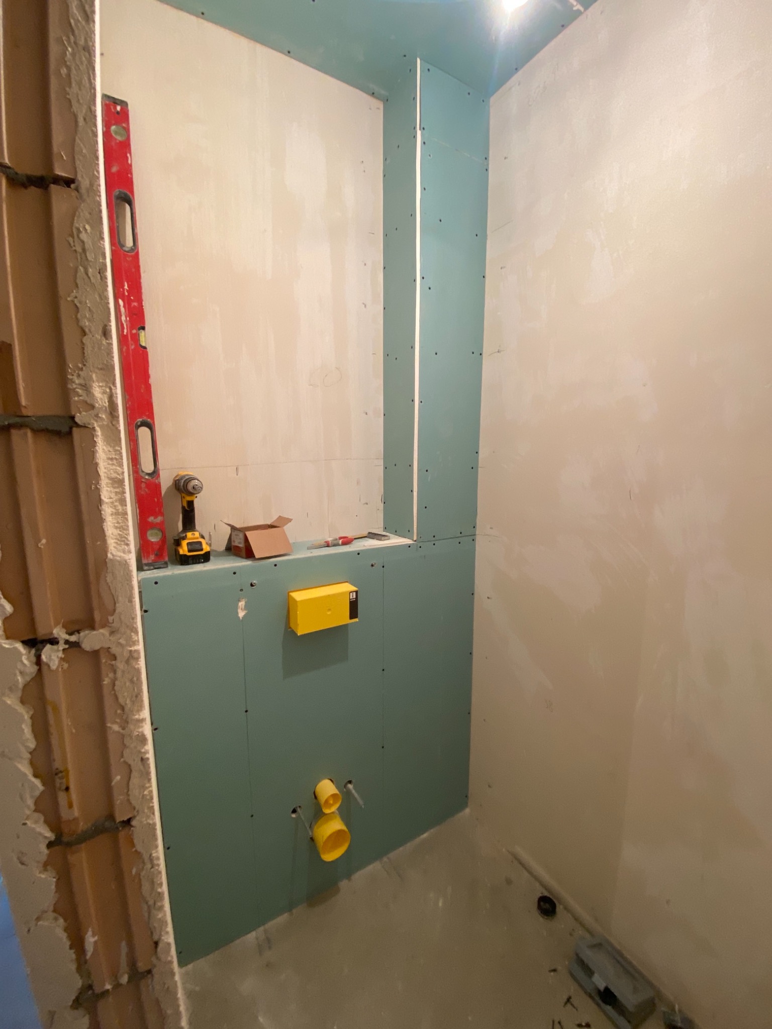 Wnętrze łazienki w trakcie remontu: widoczne płyty gipsowo-kartonowe, poziomica, wkrętarka, puszka instalacyjna i fragment odsłoniętej ściany z cegły.