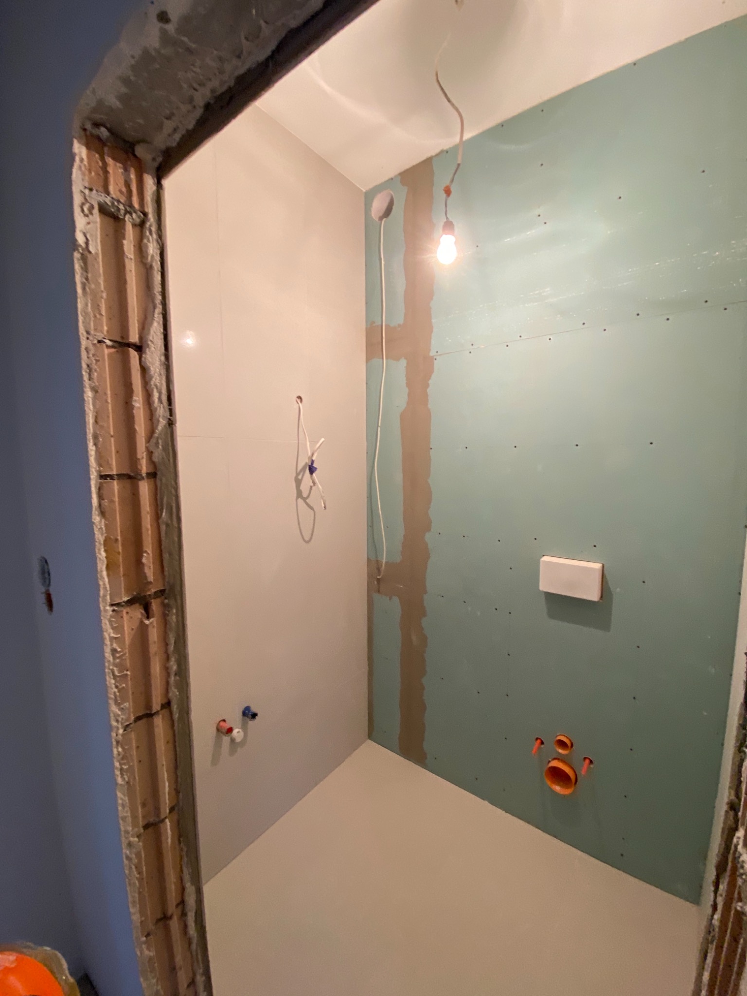 Wnętrze łazienki w trakcie remontu: ściany częściowo wyłożone białymi płytkami, częściowo pokryte zielonymi płytami gipsowo-kartonowymi, widoczne instalacje wodno-kanalizacyjne, surowy ościeżnica...