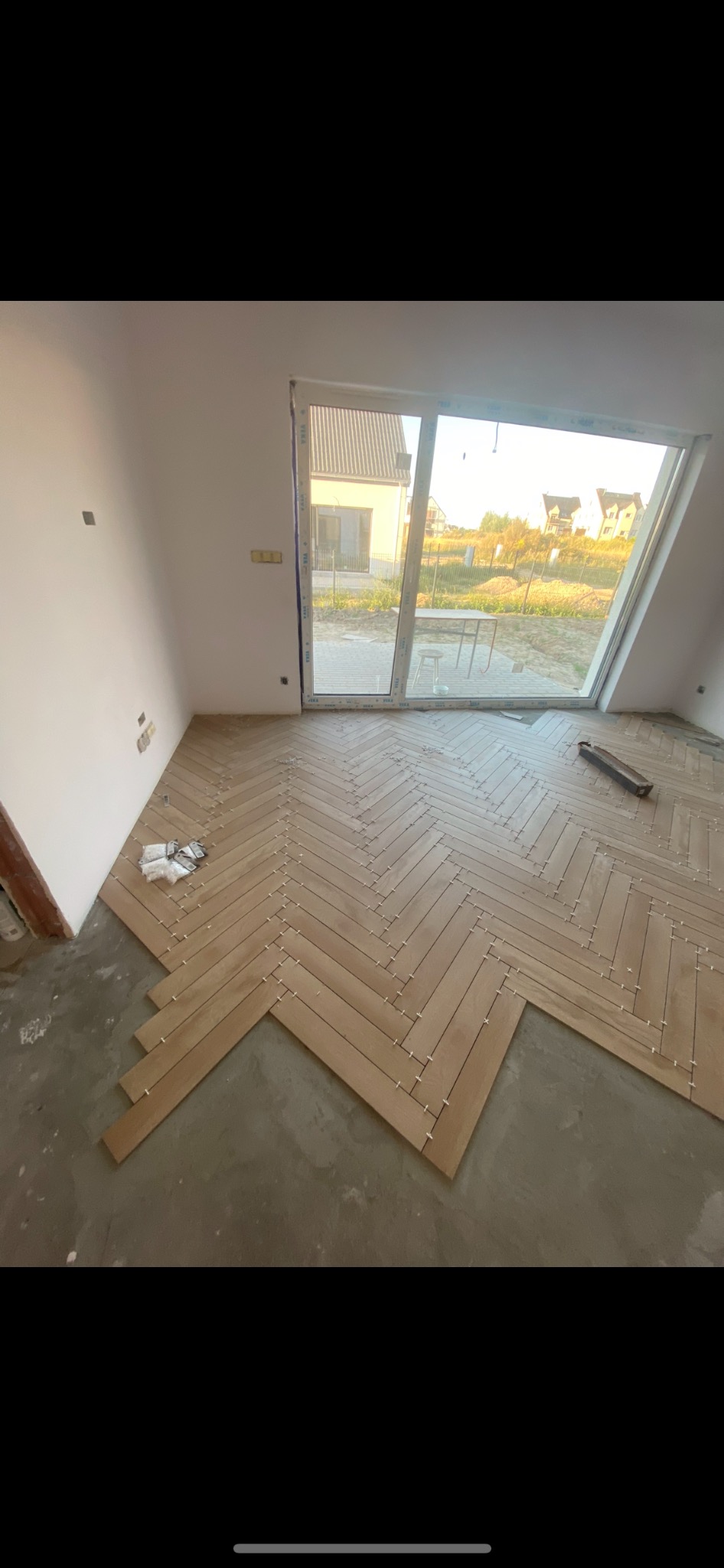 Układanie drewnianej podłogi w jodełkę francuską w jasnym wnętrzu, widoczne krzyżyki dystansowe, duże okno z widokiem na ogród i dom.