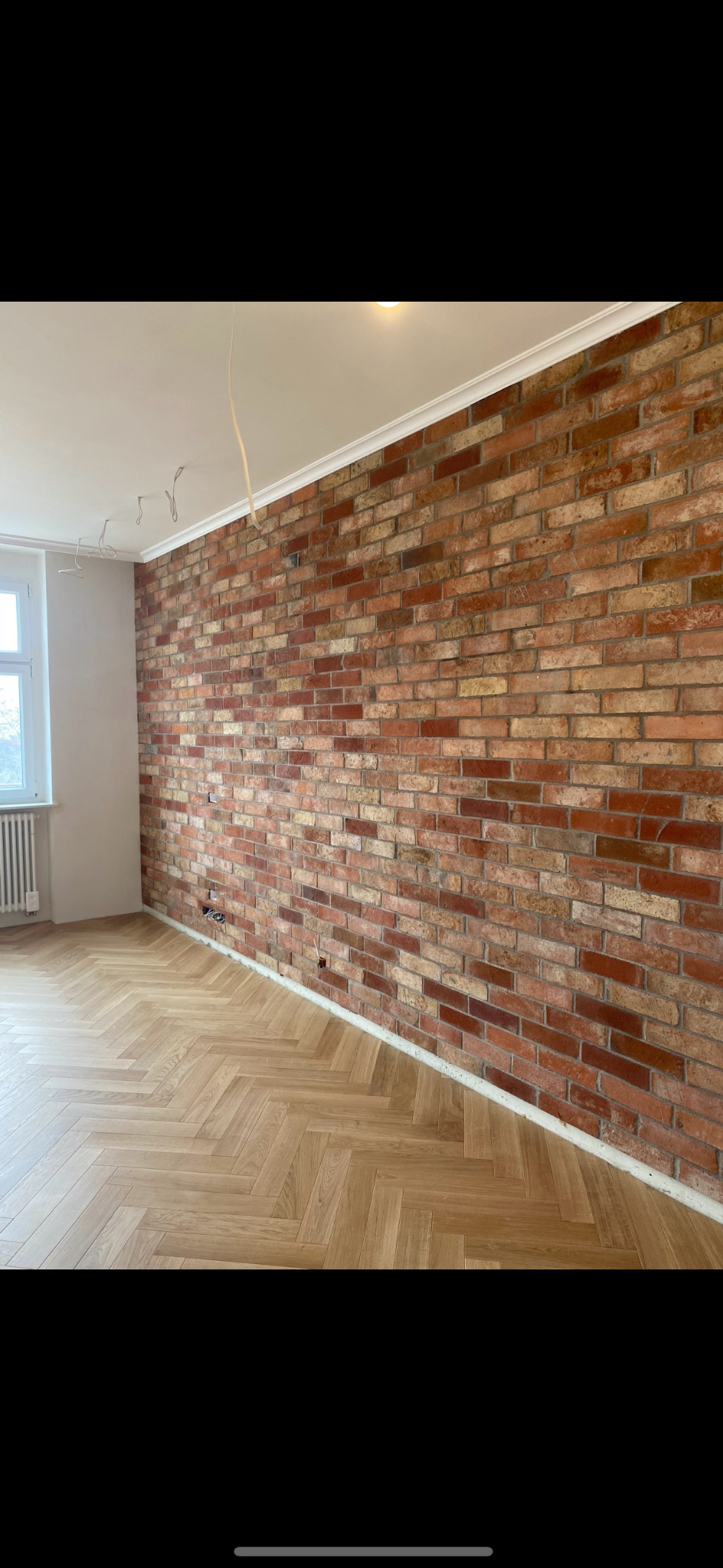 Śrem: Wykończona ściana z cegły i parkiet w jodełkę. Widoczne przewody elektryczne, białe listwy przypodłogowe i okno. Wnętrze w trakcie prac wykończeniowych.