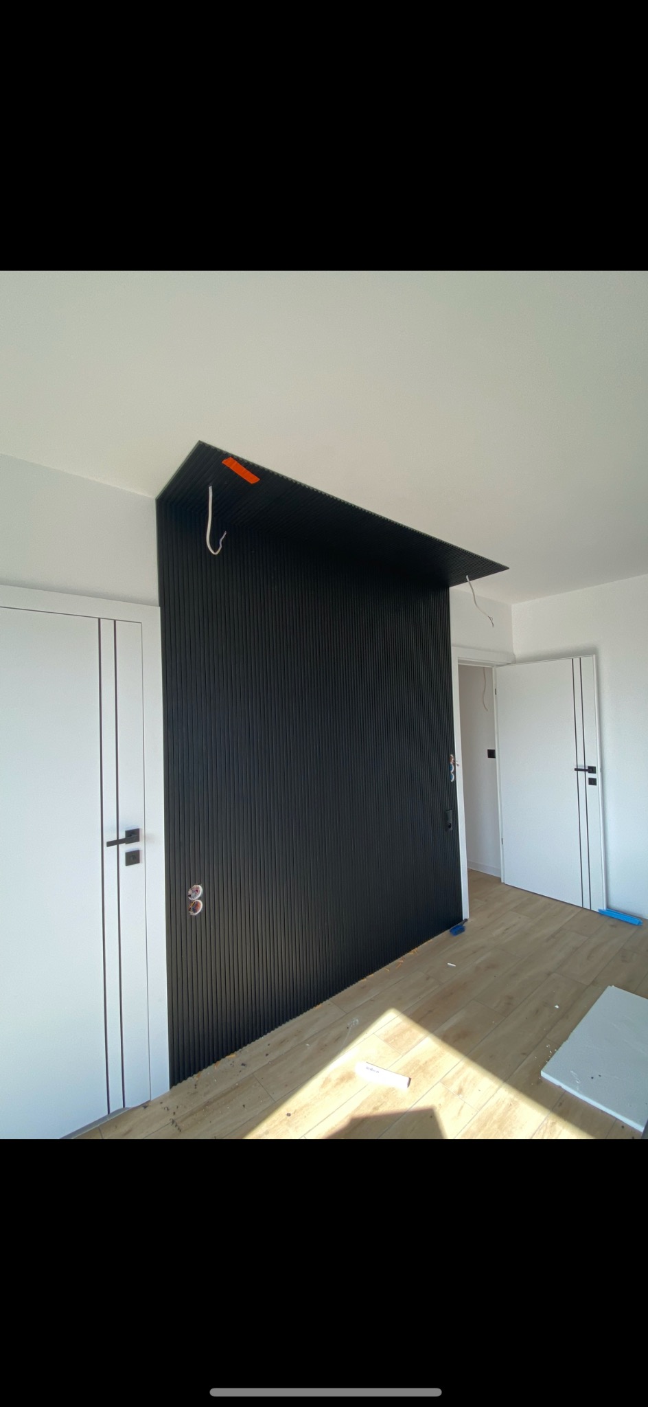 Montaż czarnej, pionowej boazerii na ścianie w pokoju z dwiema białymi drzwiami i panelami podłogowymi, widoczne kable i uchwyty montażowe.