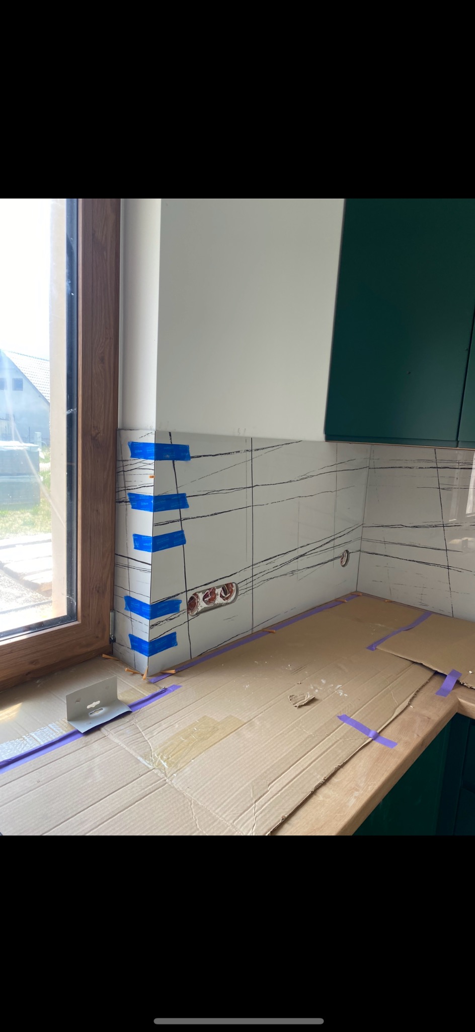 Ściana z białych płytek z czarnym wzorem, zabezpieczona niebieską taśmą, okno z drewnianą ramą i fragment zielonej szafki. Etap prac wykończeniowych.