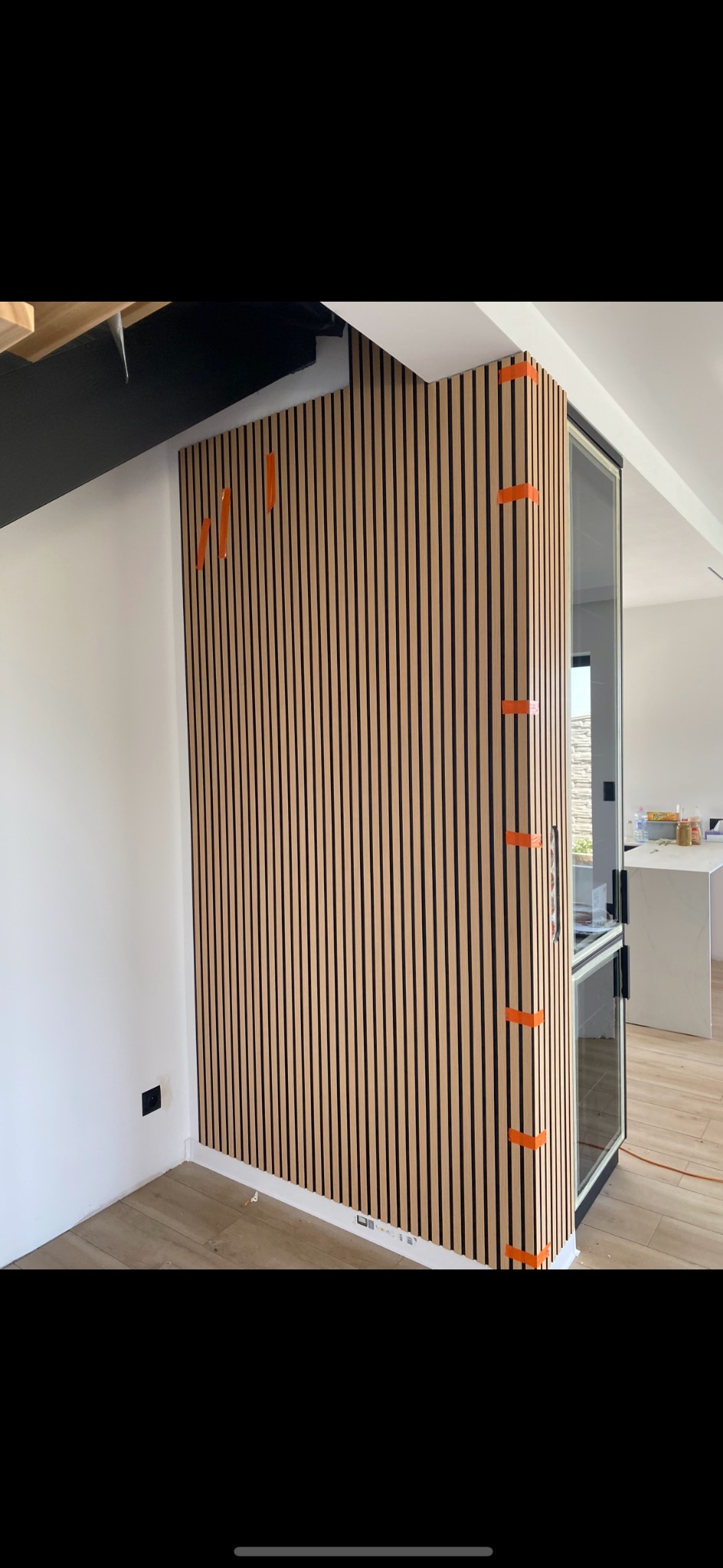 Montaż paneli lamelowych na ścianie, zabezpieczonych taśmą malarską. Widoczny fragment podłogi i narożnik ściany z oknem. Nowoczesny design wnętrza.