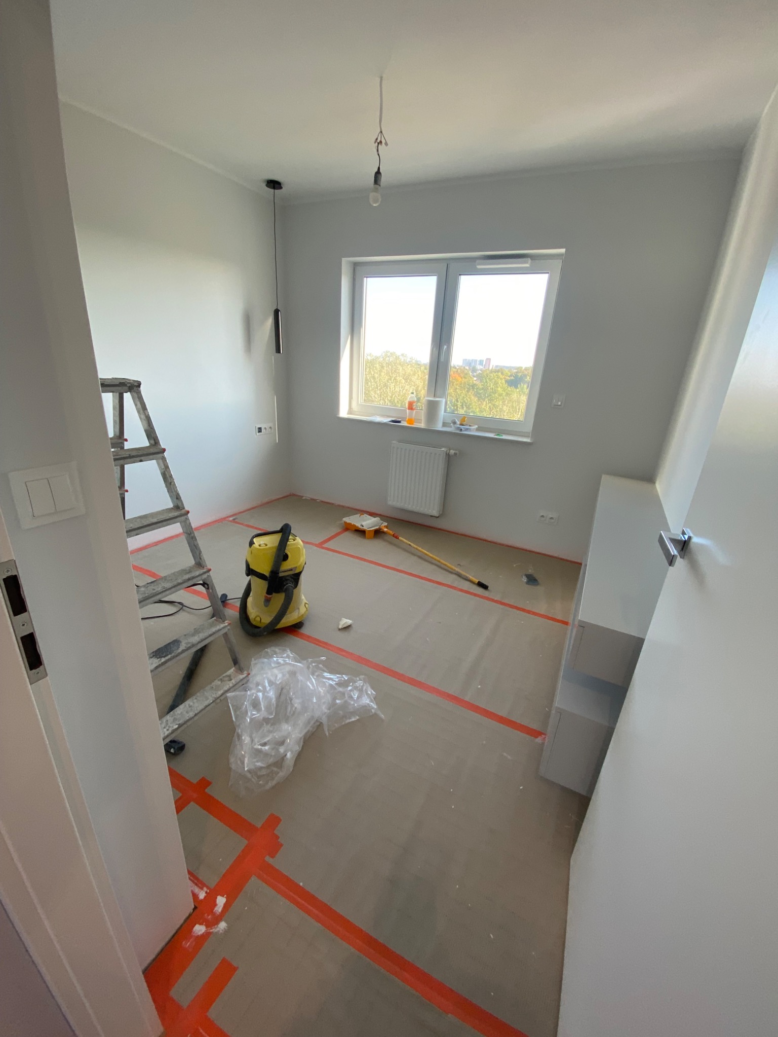 Pokój po malowaniu ścian na szaro, zabezpieczona podłoga, drabina, odkurzacz, widok zza uchylonych drzwi.