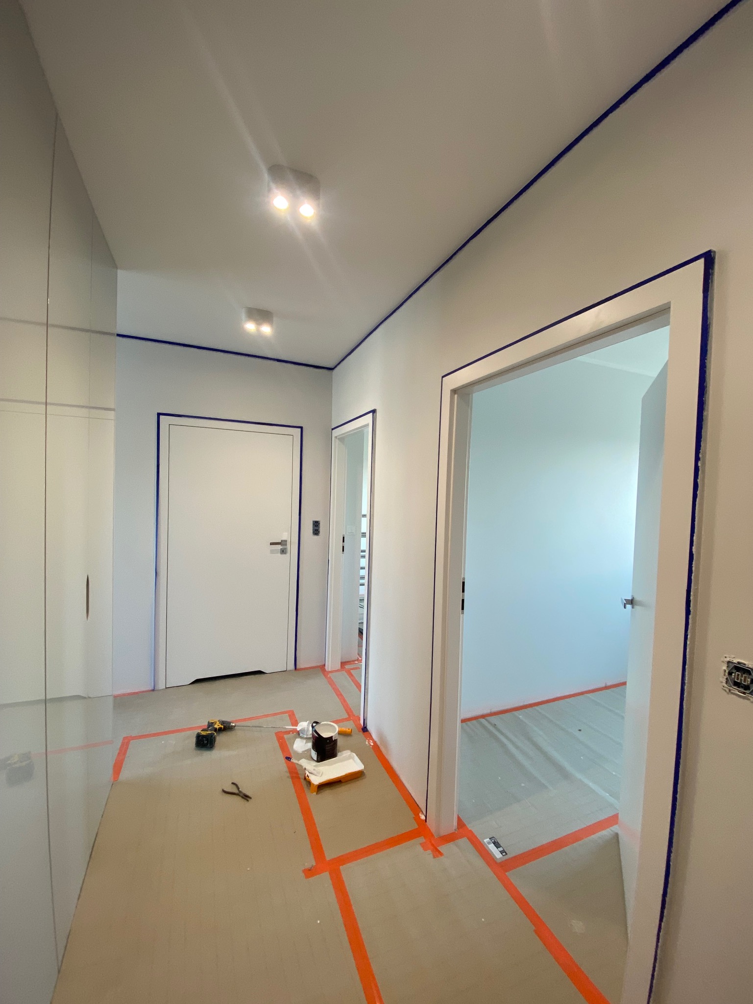 Wnętrze w trakcie prac malarskich: ściany i framugi zabezpieczone taśmą malarską, widoczne narzędzia (pędzel, wałek) i farba. Oświetlenie sufitowe, minimalistyczny design.