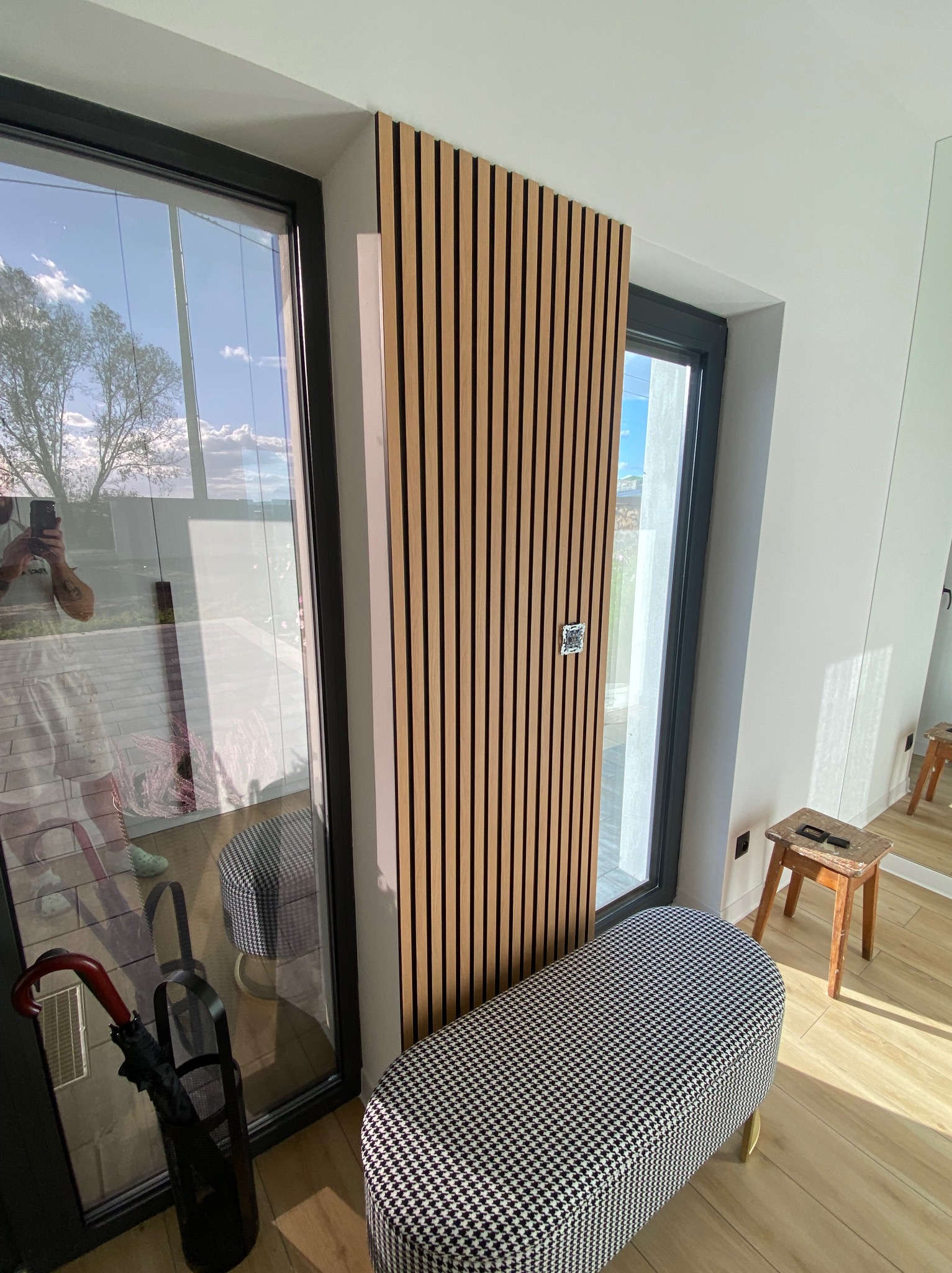 Nowoczesny przedpokój z drewnianymi lamelami na ścianie obok okna, wzorzystą ławką i drewnianym stołkiem. Widoczny fragment lustra i odbicie osoby robiącej zdjęcie.