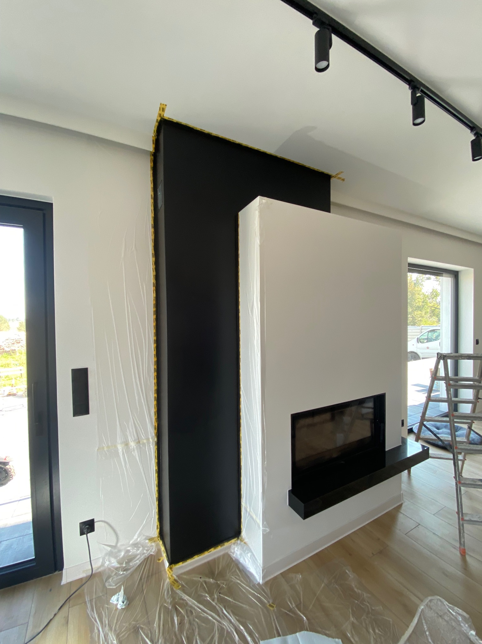 Wnętrze w trakcie malowania: fragment ściany z czarnym elementem zabezpieczonym taśmą, kominek i drabina. Folia ochronna na podłodze, światło punktowe na suficie.