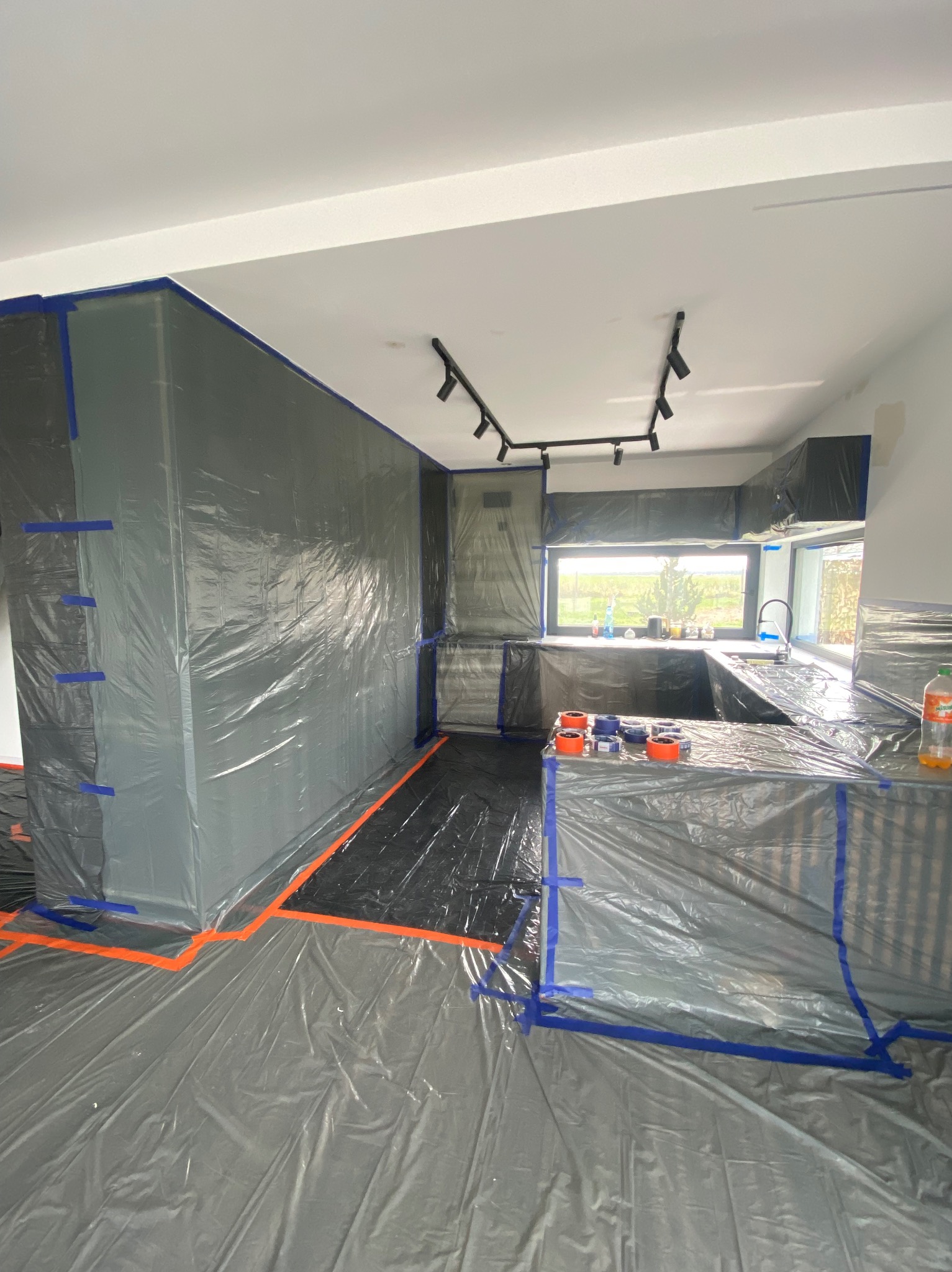 Kuchnia zabezpieczona folią i taśmą malarską przed malowaniem. Widoczne okno, szafki kuchenne i podłoga przygotowane do prac remontowych. Oświetlenie sufitowe.