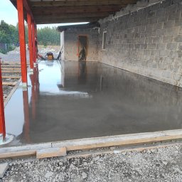 Floorwit Vitalii Mezhduhornyi - Świeżo wylana, gładka, szara posadzka w trakcie realizacji, widoczne czerwone drewniane słupy podtrzymujące zadaszenie, szalunki i gruz na obrzeżach.