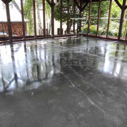 Floorwit Vitalii Mezhduhornyi - Świeżo wylana, mokra posadzka w zadaszonej konstrukcji z drewnianymi słupami, odbijająca otoczenie, z widocznymi śladami po narzędziach i drobnymi zabrudzeniami.