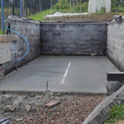 Floorwit Vitalii Mezhduhornyi - Świeżo wylana, szara posadzka w zagłębieniu otoczonym murem z bloczków, widoczny fragment granicy z grysem i elementami obrzeża.