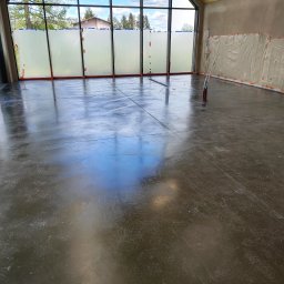 Floorwit Vitalii Mezhduhornyi - Świeżo wylana, błyszcząca, ciemnoszara posadzka w przestronnym pomieszczeniu z dużymi oknami zabezpieczonymi folią malarską, widoczne narzędzia i ślady stóp.