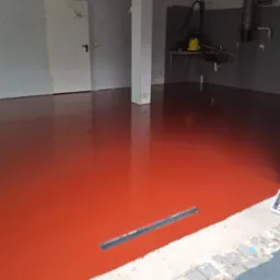 Błyszcząca, czerwona posadzka żywiczna w garażu z odpływem liniowym. Widoczne wiadro z farbą i narzędzia. Metalowy sufit i białe ściany w tle.