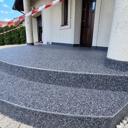Floorwit Vitalii Mezhduhornyi - Wykończone schody wejściowe z szarej, drobnoziarnistej posadzki żywicznej, zabezpieczone taśmą ostrzegawczą. Widoczne kolumny i fragment elewacji budynku.