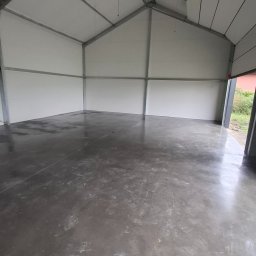 Floorwit Vitalii Mezhduhornyi - Wnętrze hali z szarą, gładką posadzką i białymi ścianami. Widoczna metalowa konstrukcja dachu i częściowo otwarte drzwi w tle. Ślady na posadzce.