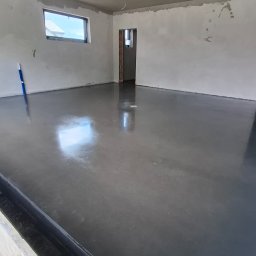 Floorwit Vitalii Mezhduhornyi - Nowo wylana, lśniąca, ciemnoszara posadzka w przestronnym, nieukończonym pomieszczeniu z oknem i otwartym przejściem, z widocznymi przewodami elektrycznymi zwisającymi z sufitu.