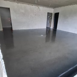 Floorwit Vitalii Mezhduhornyi - Nowo wylana, ciemnoszara, błyszcząca wylewka podłogowa w pustym pomieszczeniu z niedokończonymi ścianami i zwisającymi przewodami z sufitu.