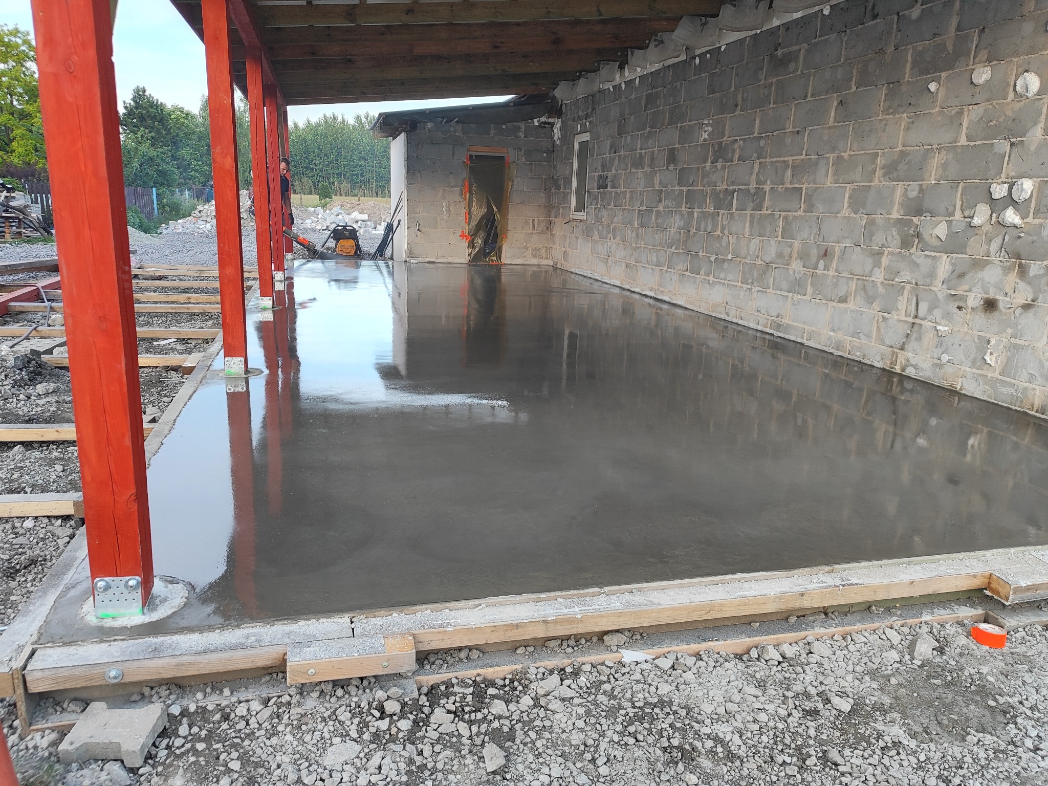 Świeżo wylana, gładka, szara posadzka w trakcie realizacji, widoczne czerwone drewniane słupy podtrzymujące zadaszenie, szalunki i gruz na obrzeżach.