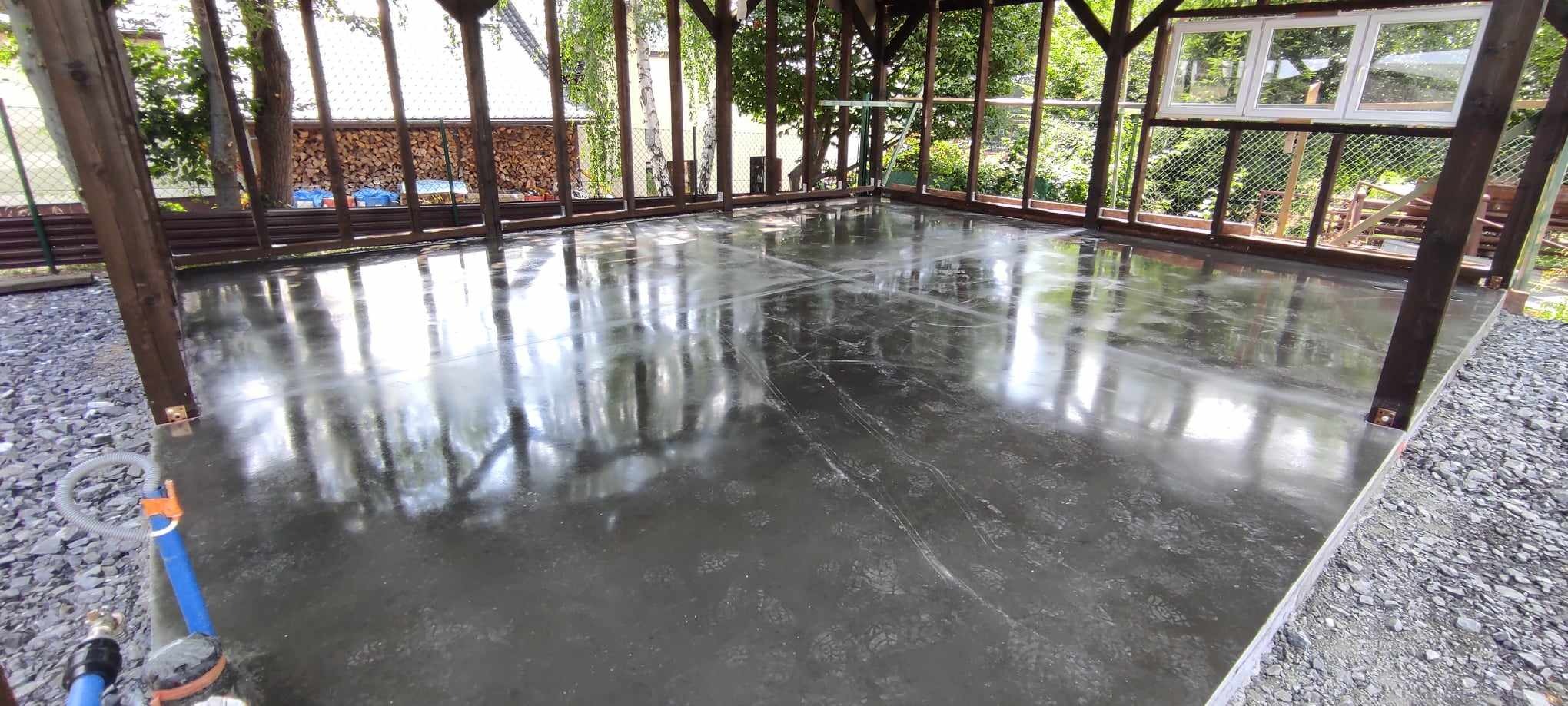 Świeżo wylana, mokra posadzka w zadaszonej konstrukcji z drewnianymi słupami, odbijająca otoczenie, z widocznymi śladami po narzędziach i drobnymi zabrudzeniami.
