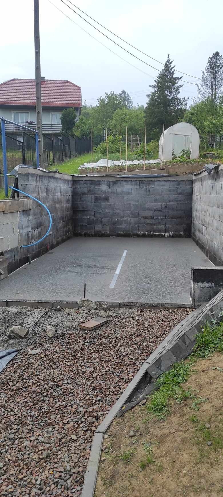 Świeżo wylana, szara posadzka w zagłębieniu otoczonym murem z bloczków, widoczny fragment granicy z grysem i elementami obrzeża.