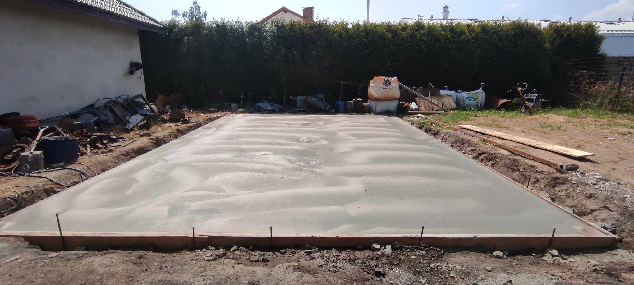 Świeżo wylana, mokra betonowa posadzka przemysłowa w drewnianym szalunku, na zewnątrz, z widocznymi workami zaprawy w tle.