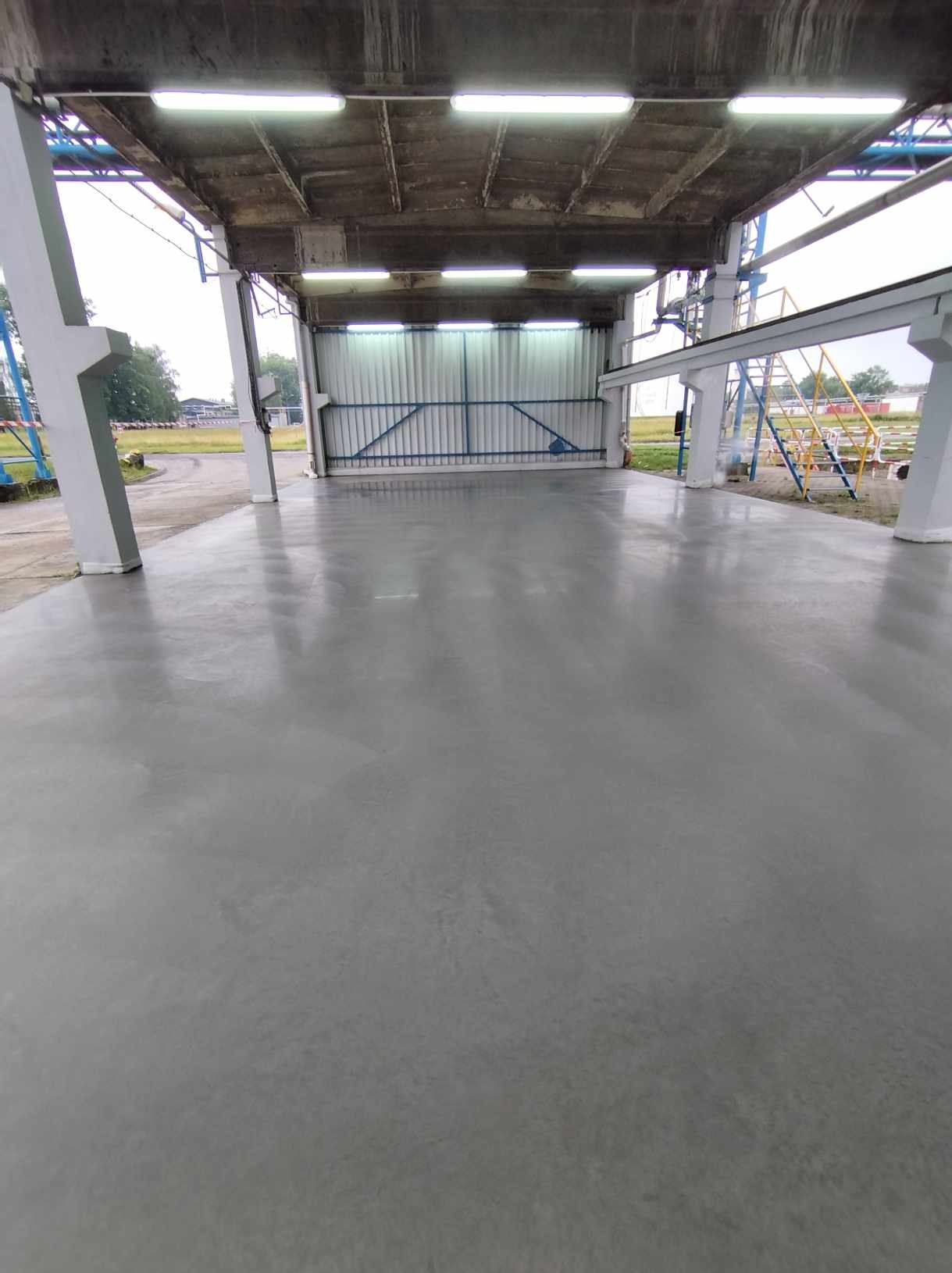 Gładka, szara posadzka w hali przemysłowej z betonowym sufitem i metalową bramą w tle, oświetlona lampami jarzeniowymi.