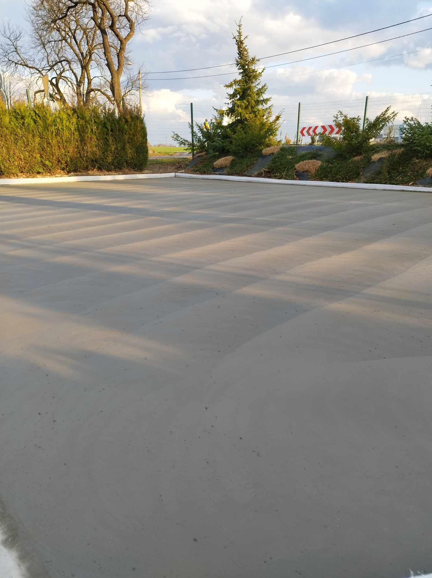 Świeżo wylana, gładka, betonowa powierzchnia zewnętrzna z widocznymi śladami po wygładzaniu, otoczona zielenią i ogrodzeniem.