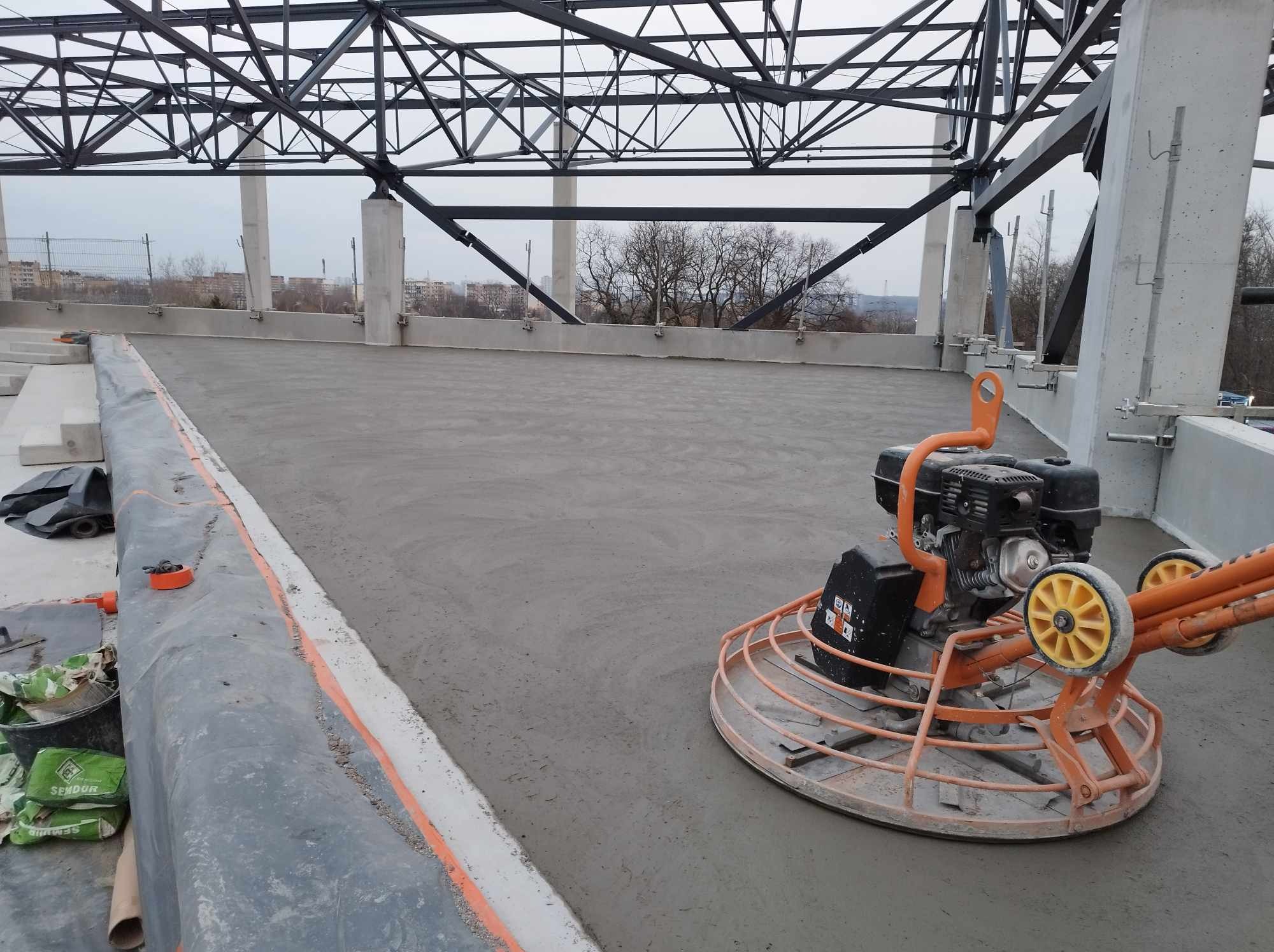 Wyrównywanie świeżo wylanej posadzki przemysłowej na budowie, widoczna zacieraczka mechaniczna i worki z zaprawą w tle, konstrukcja stalowa dachu.