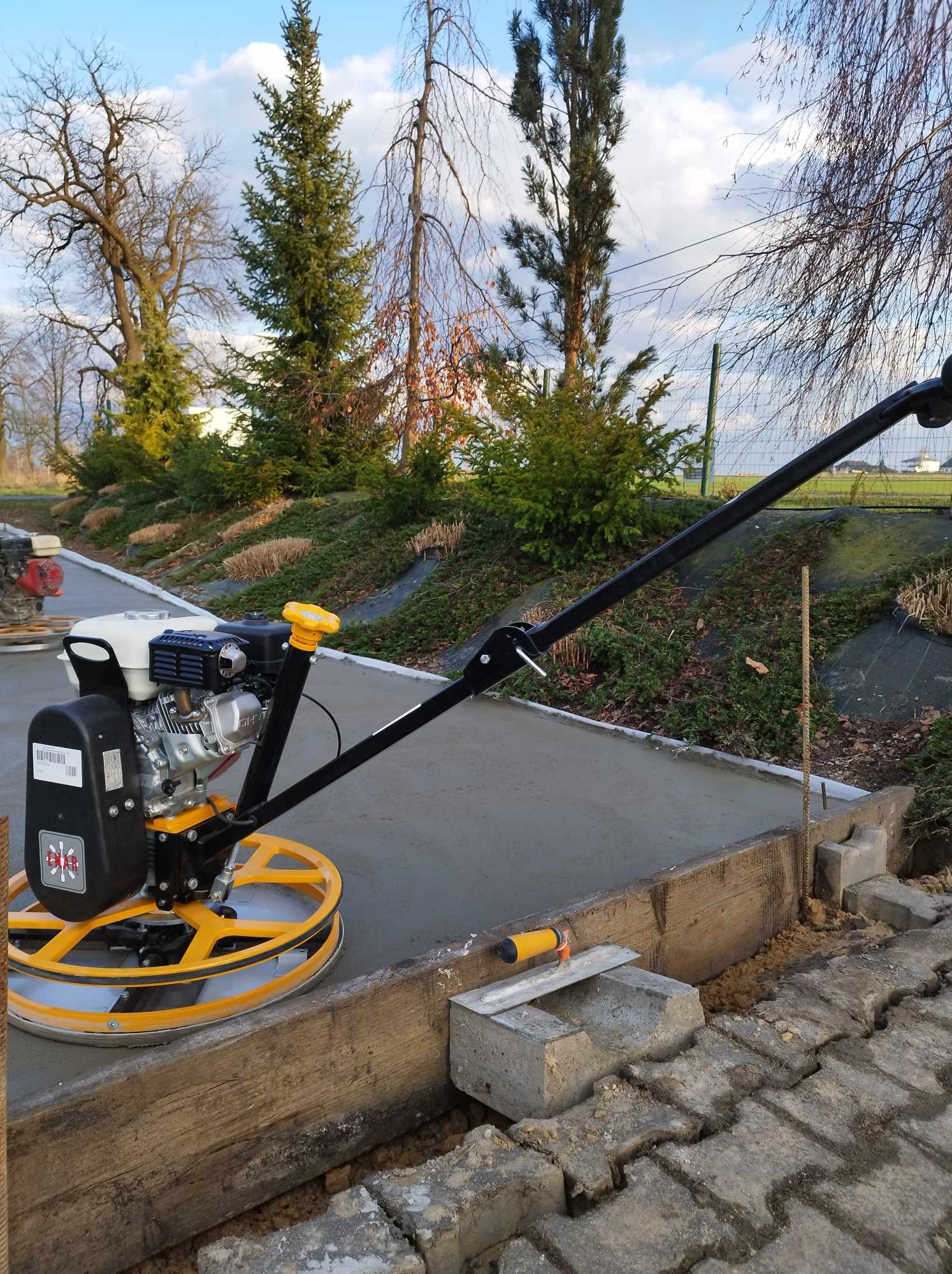 Zacieraczka mechaniczna z żółtymi elementami podczas pracy na wylewce betonowej, widoczne drewniane szalunki i kostka brukowa w tle.