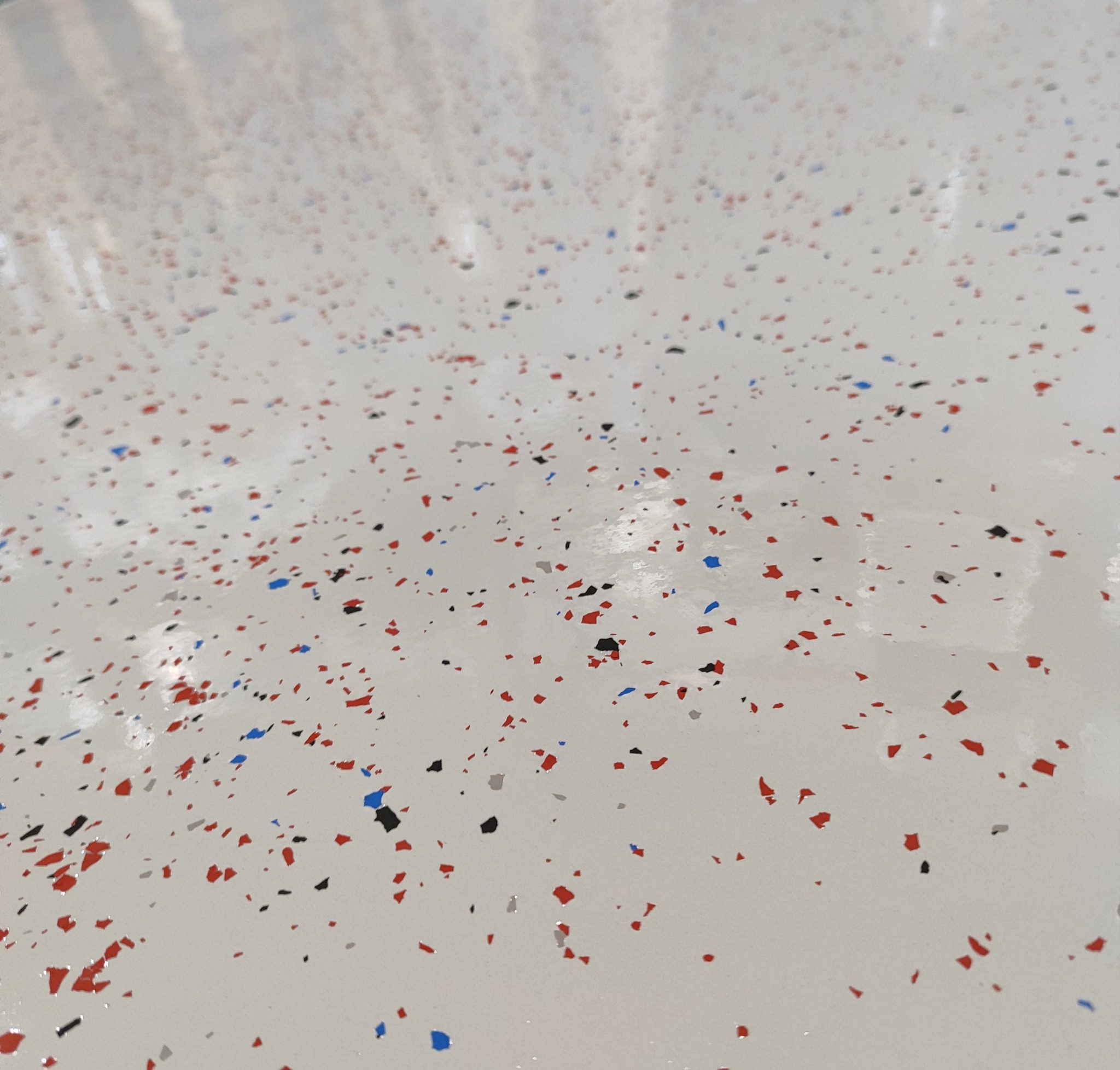 Gładka, jasna posadzka z żywicy z wtopionymi kolorowymi płatkami (czerwonymi, niebieskimi, czarnymi i szarymi) o nieregularnym kształcie, tworzącymi dekoracyjny wzór.