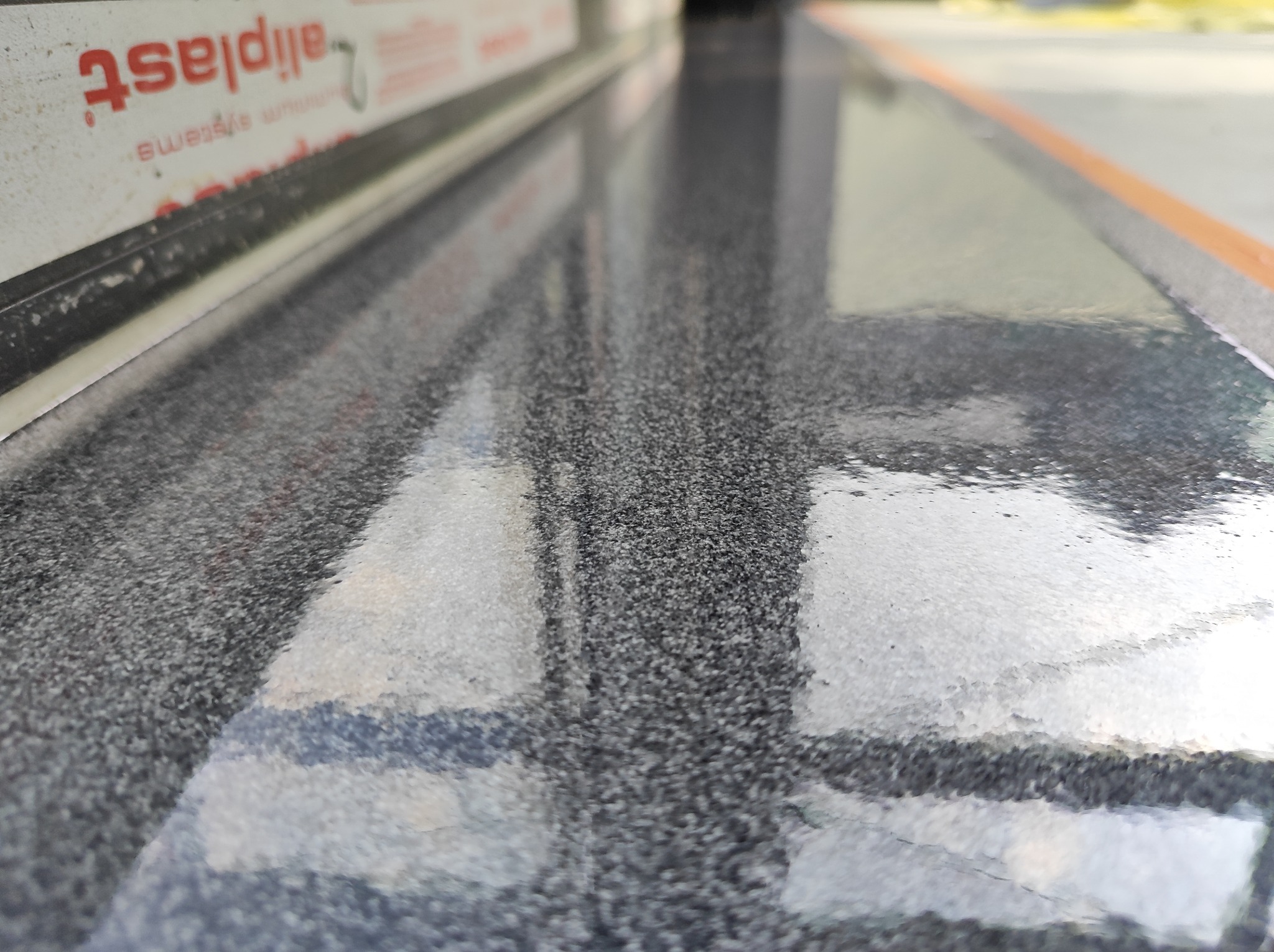 Błyszcząca, szara posadzka z widocznym odbiciem okna i fragmentem ramy okiennej z napisem 'aliplast'.