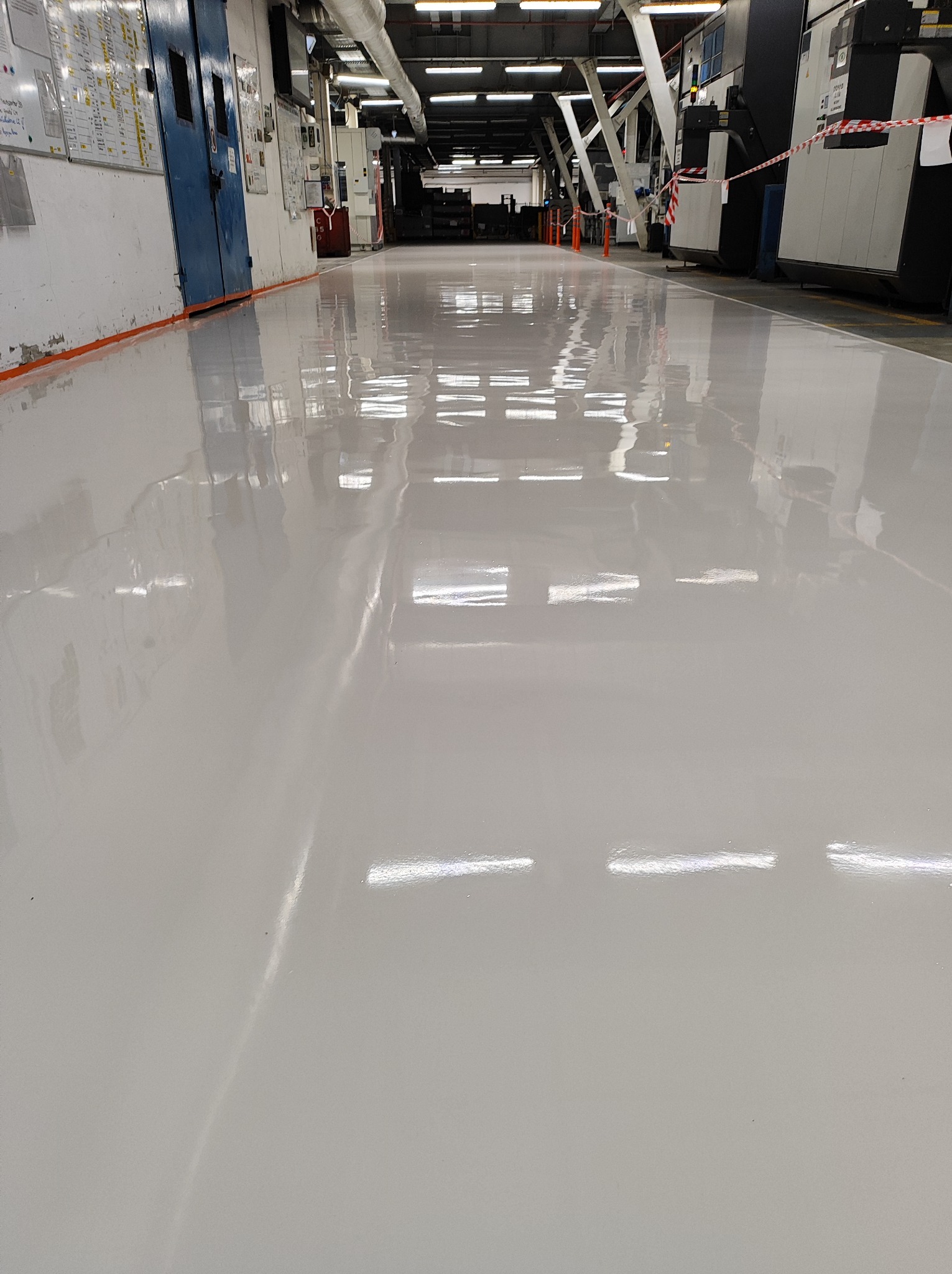 Błyszcząca, jasnoszara posadzka w hali produkcyjnej z widocznymi maszynami i instalacjami pod sufitem.