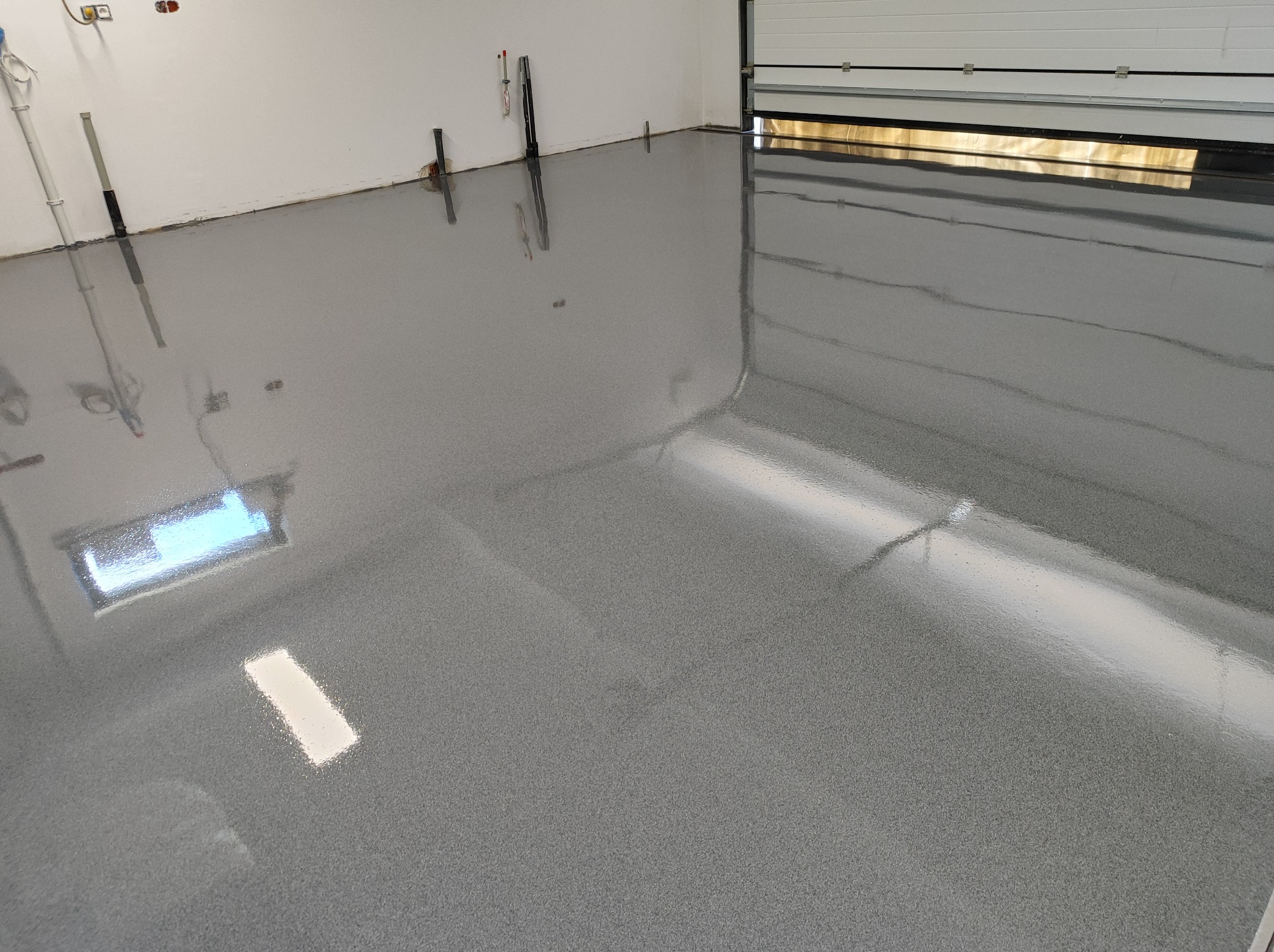 Świeżo wylana, błyszcząca, szara posadzka w garażu z widocznymi instalacjami na ścianie i otwartą bramą garażową w tle.