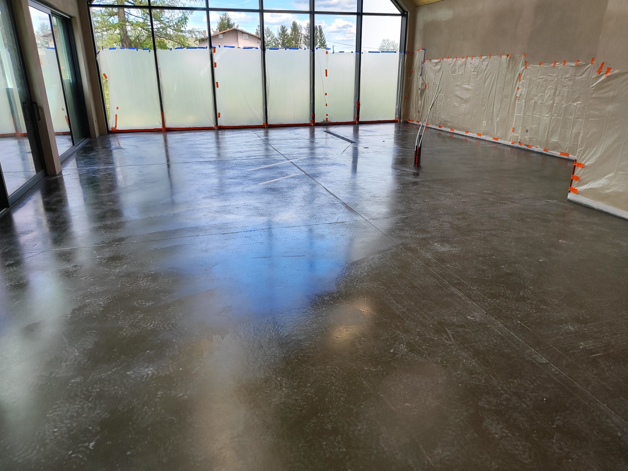 Świeżo wylana, błyszcząca, ciemnoszara posadzka w przestronnym pomieszczeniu z dużymi oknami zabezpieczonymi folią malarską, widoczne narzędzia i ślady stóp.
