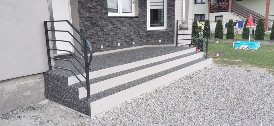 Zewnętrzne schody wejściowe z czarno-białą, kamyczkową nawierzchnią i czarną metalową balustradą, prowadzące do domu z elewacją z szarego kamienia.