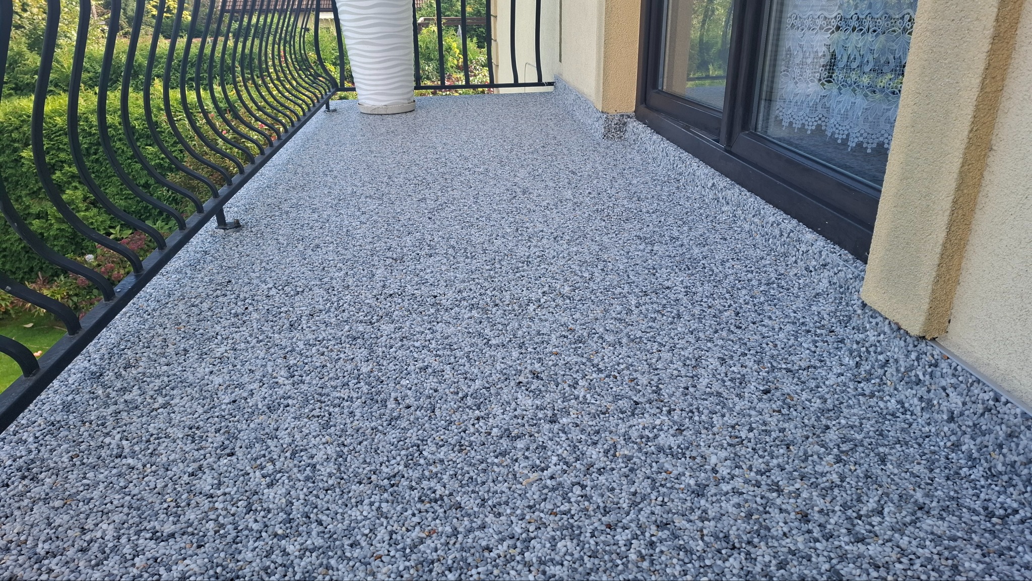 Balkon z szarą, kamyczkową posadzką żywiczną, widok z góry na balustradę z kutego żelaza i roślinność w tle.