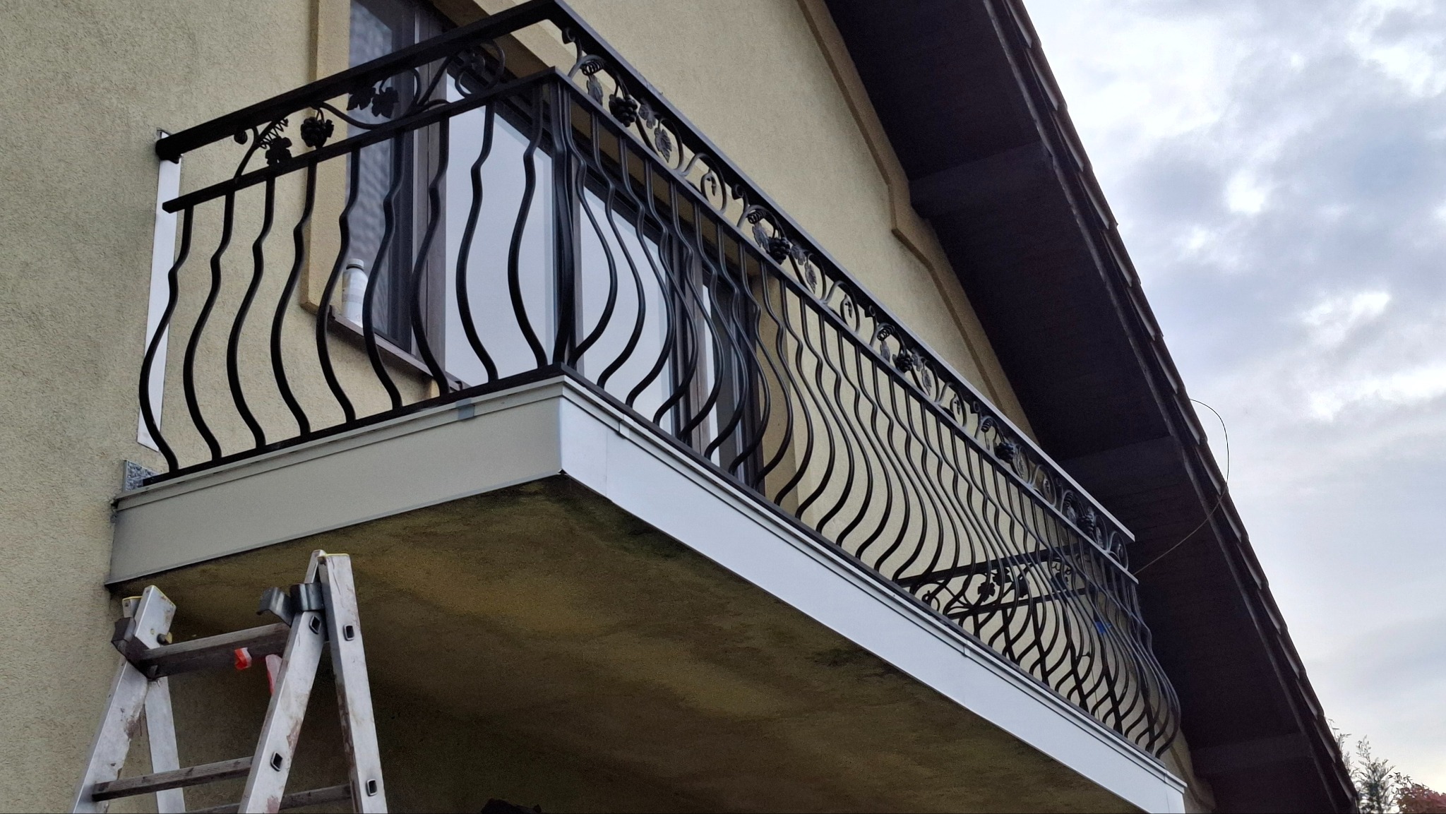 Balkon z czarną, ozdobną balustradą z motywem winogron, widok z dołu, drabina oparta o ścianę budynku.