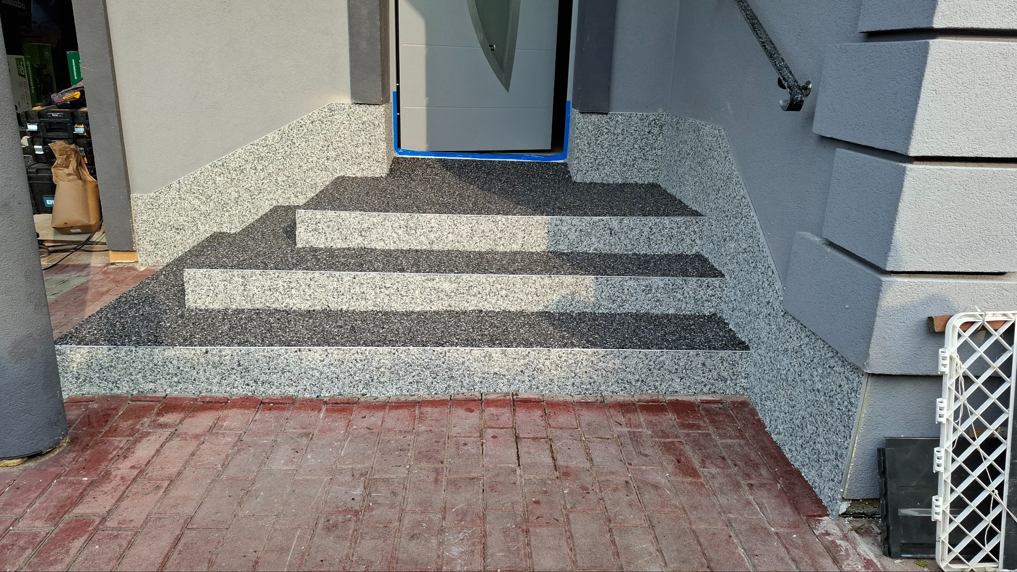 Granitowe schody wejściowe z dwukolorową powierzchnią, zabezpieczone niebieską taśmą malarską przy progu drzwi, z widoczną czerwoną kostką brukową przed nimi.