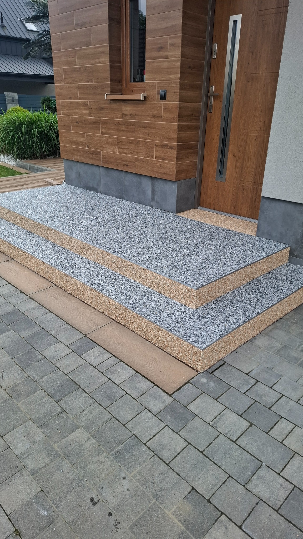 Zewnętrzne schody wejściowe wykończone szarą posadzką żywiczną z widoczną strukturą kruszywa, prowadzące do drzwi wejściowych domu z drewnianą elewacją.