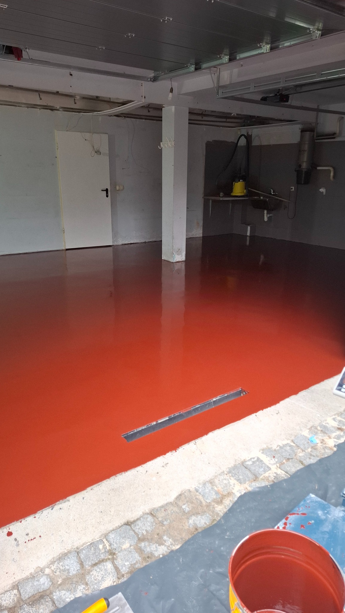 Błyszcząca, czerwona posadzka żywiczna w garażu z odpływem liniowym. Widoczne wiadro z farbą i narzędzia. Metalowy sufit i białe ściany w tle.