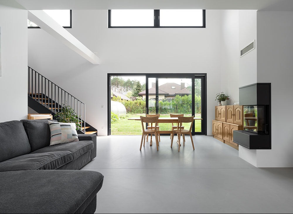 Jasne wnętrze z szarą posadzką, minimalistycznymi schodami i dużym oknem na ogród. Widoczny fragment sofy i kominek. Nowoczesny design z naturalnym światłem.