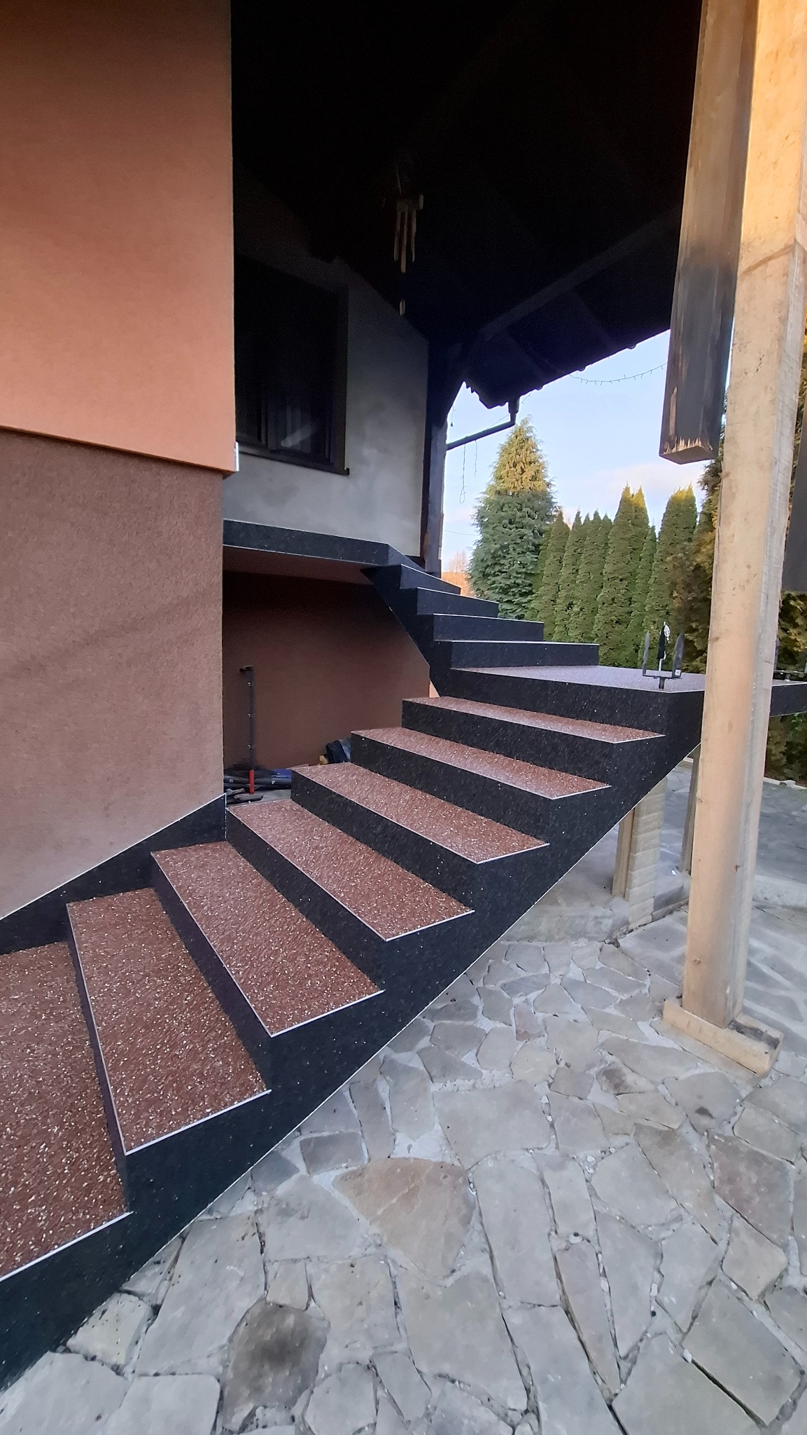 Zewnętrzne schody z brązową, żywiczną nawierzchnią i czarnymi bokami, prowadzące do domu. Widoczna kamienna posadzka i filar podtrzymujący zadaszenie.