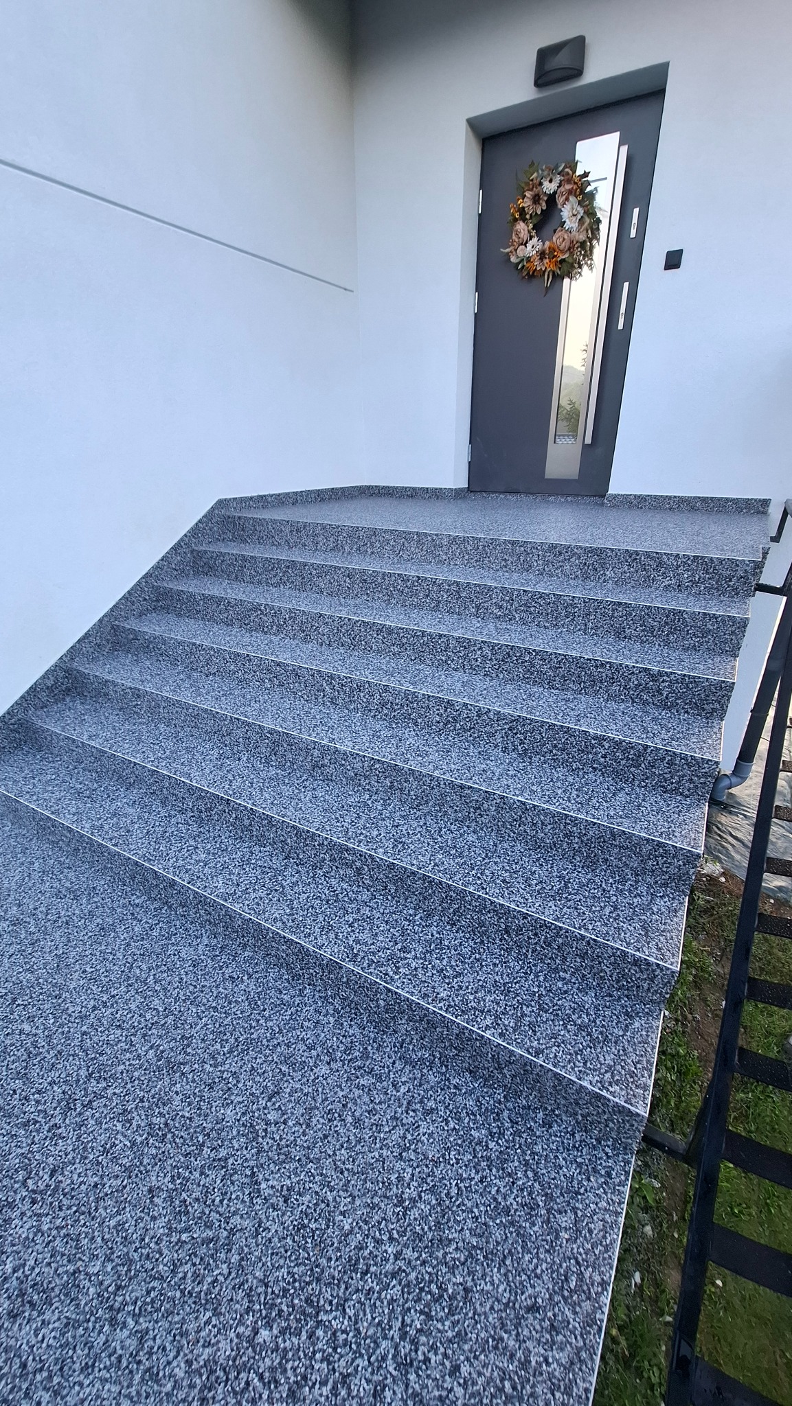 Zewnętrzne schody wejściowe wykończone szarą, żywiczną posadzką z drobnym kruszywem, prowadzące do nowoczesnych drzwi z wieńcem. Minimalistyczny design.