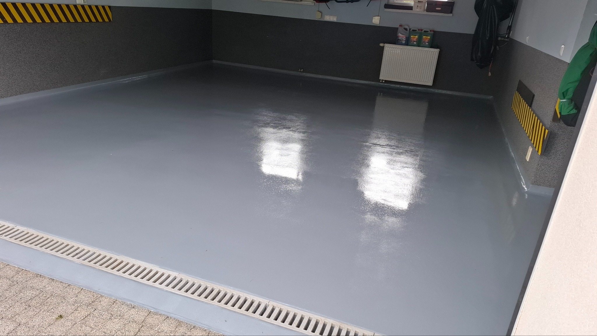 Szara, gładka posadzka w garażu z odpływem liniowym. Widoczne żółto-czarne pasy ostrzegawcze na ścianie i grzejnik. Błyszcząca powierzchnia odbija światło.