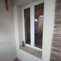 G & G ADAM GÓRKA - Białe okno PCV na balkonie z widokiem na blok mieszkalny, obok okna stoi mała lampka naftowa, po prawej stronie widoczna bambusowa roleta.