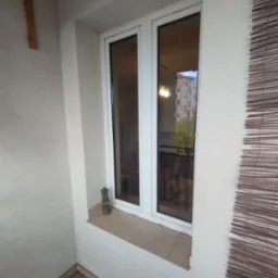 Białe okno PCV na balkonie z widokiem na blok mieszkalny, obok okna stoi mała lampka naftowa, po prawej stronie widoczna bambusowa roleta.