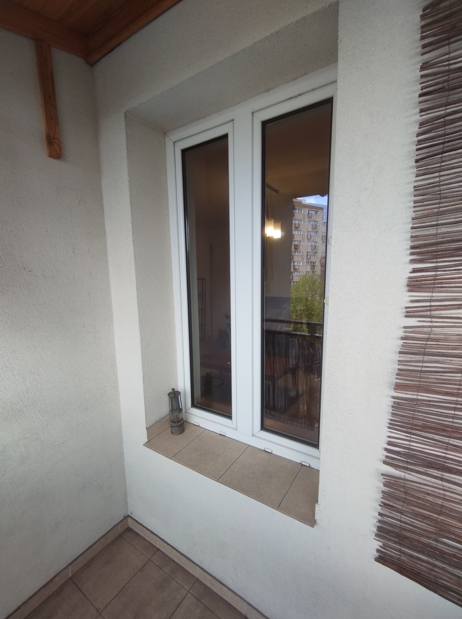 Białe okno PCV na balkonie z widokiem na blok mieszkalny, obok okna stoi mała lampka naftowa, po prawej stronie widoczna bambusowa roleta.