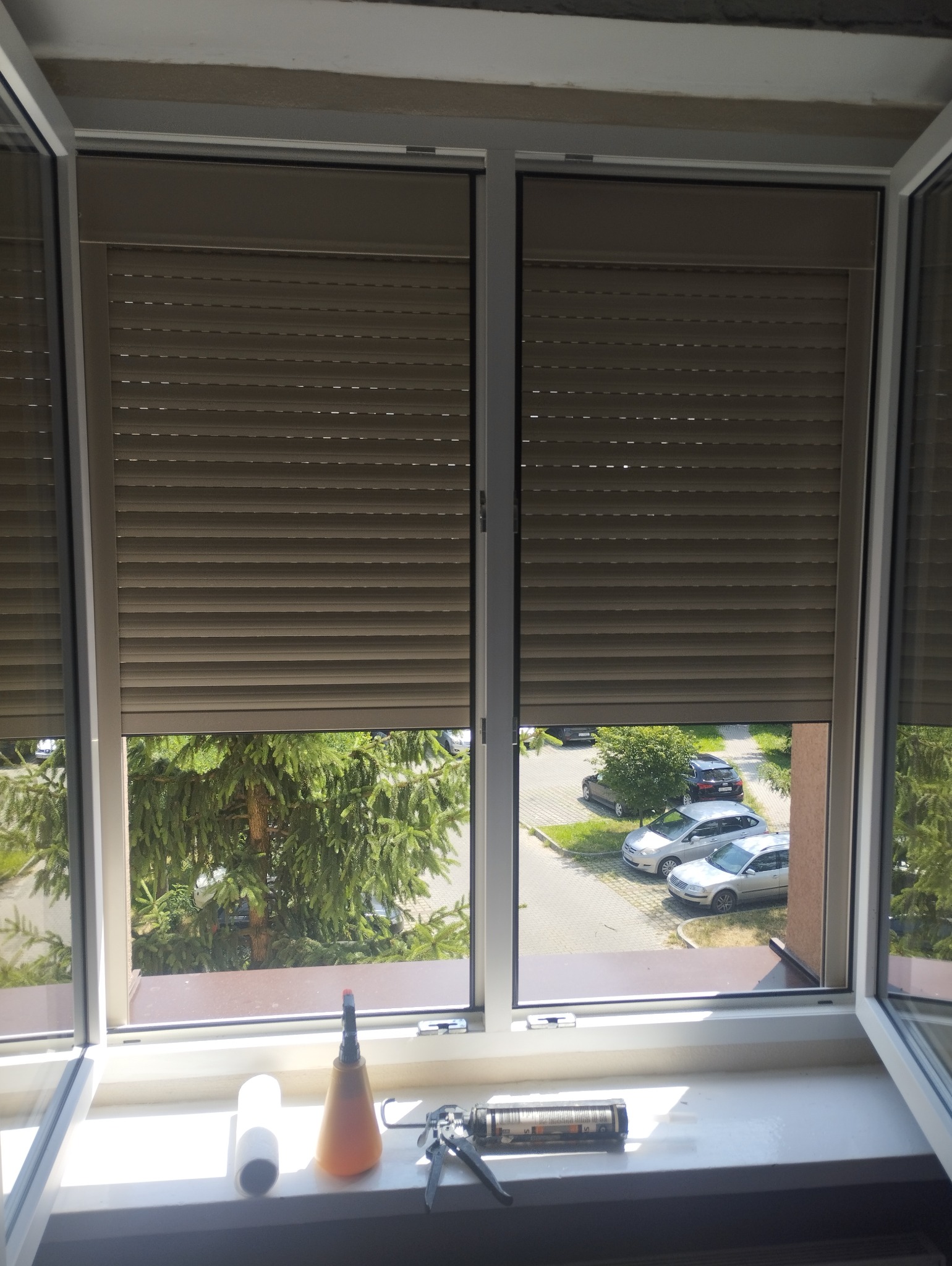 Dwa okna z częściowo opuszczonymi roletami zewnętrznymi, widok na zewnątrz z drzewami i samochodami, na parapecie narzędzia: silikon, pistolet do silikonu i rolka papieru.