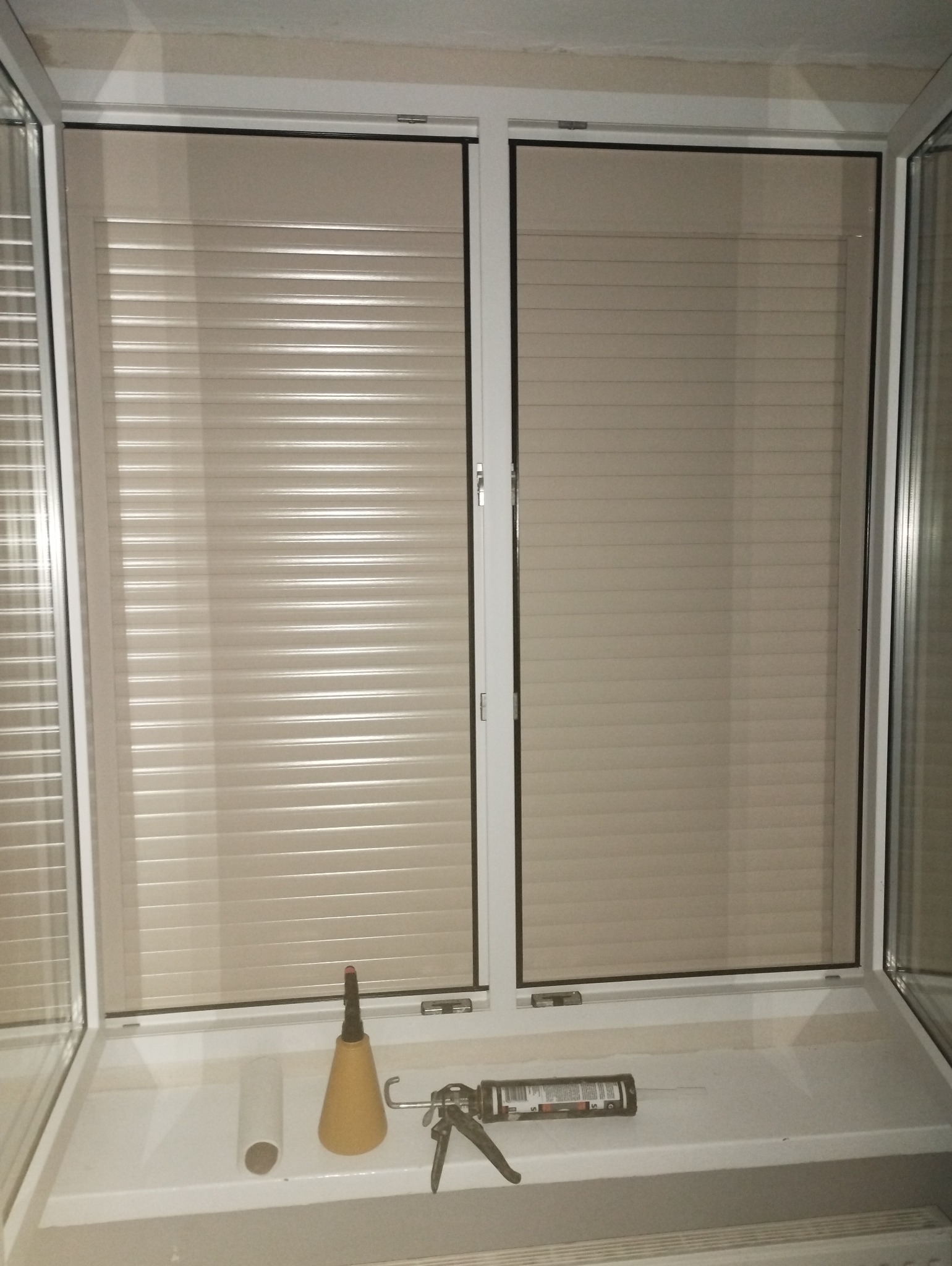 Białe okno PCV z beżowymi roletami zewnętrznymi, na parapecie leżą narzędzia: pistolet do silikonu, tuba silikonu i aplikator.